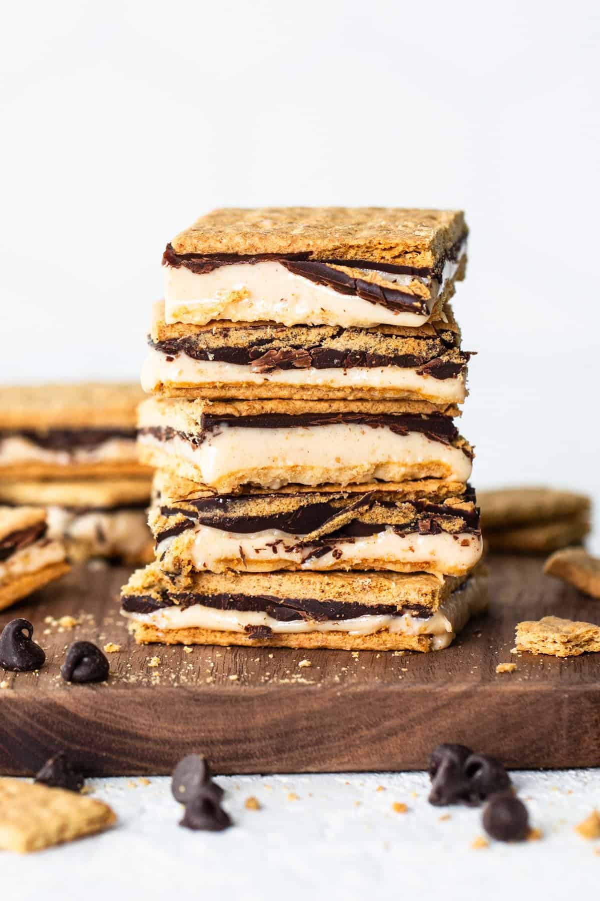 A stack of s'mores bars with layers of graham crackers, melted marshmallow, and chocolate on a wooden board, with scattered chocolate chips around.