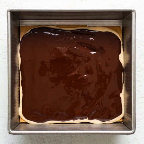 A square baking dish containing a layer of brownie topped with a glossy layer of chocolate ganache.