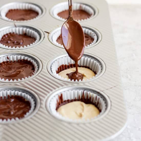 Drizzling chocolate over peanut butter in lined muffin cups using a spoon.