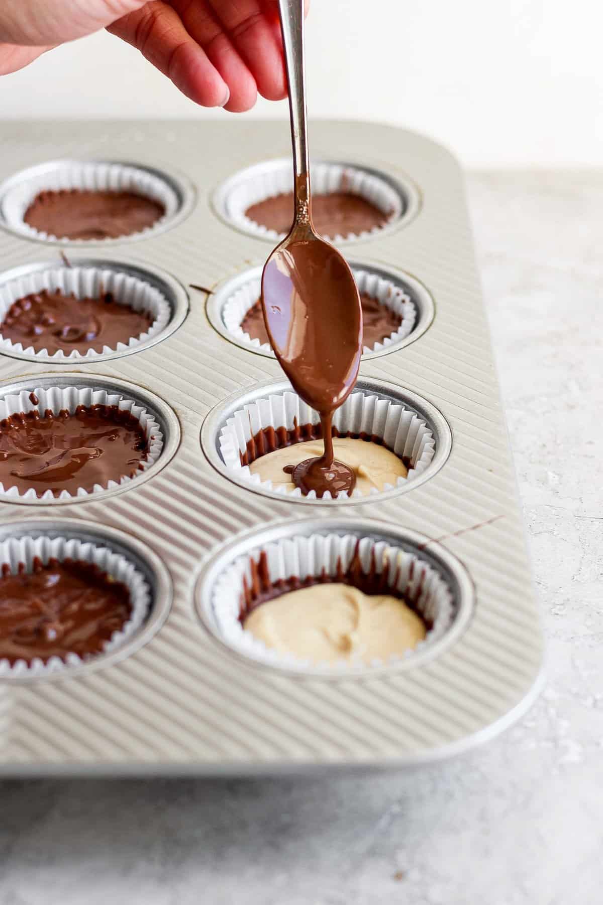 Drizzling chocolate over peanut butter in lined muffin cups using a spoon.