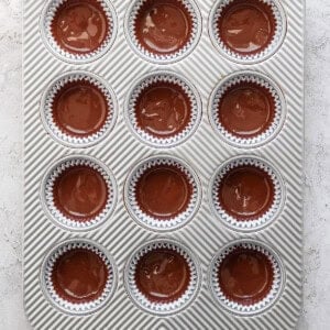 Tray of twelve chocolate cupcakes with glossy tops, each in a striped paper liner, arranged in a gray tray with a diagonal stripe pattern.