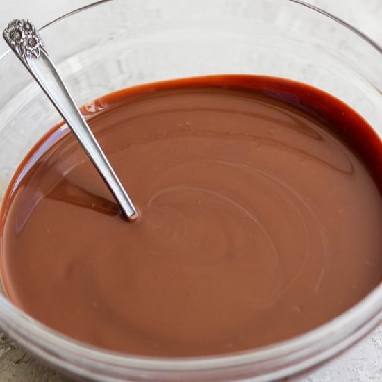 A glass bowl containing smooth melted chocolate with a metal spoon resting inside, set on a light-colored surface.