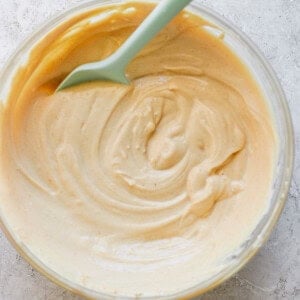 A glass bowl containing creamy peanut butter with a green spoon, photographed from overhead on a textured white surface.