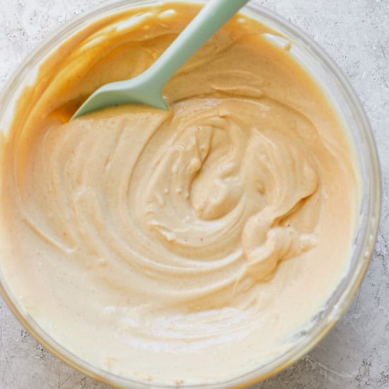 A glass bowl containing creamy peanut butter with a green spoon, photographed from overhead on a textured white surface.