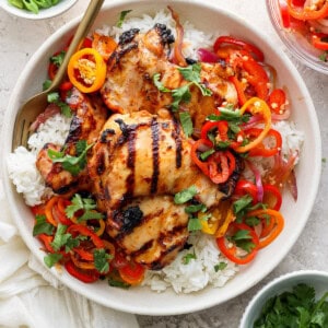 Grilled chicken thighs on a bed of white rice, garnished with sliced bell peppers, onions, and herbs in a bowl, with chopsticks on the side.