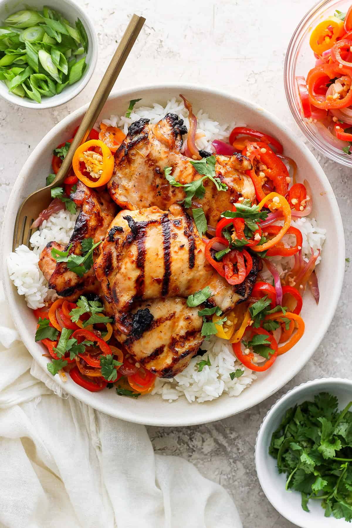 Grilled chicken thighs on a bed of white rice, garnished with sliced bell peppers, onions, and herbs in a bowl, with chopsticks on the side.