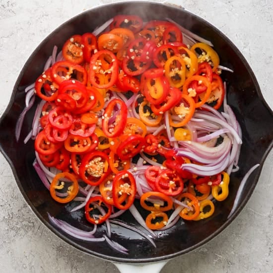 Sliced red and yellow bell peppers with red onions in a white le creuset skillet on a textured surface.