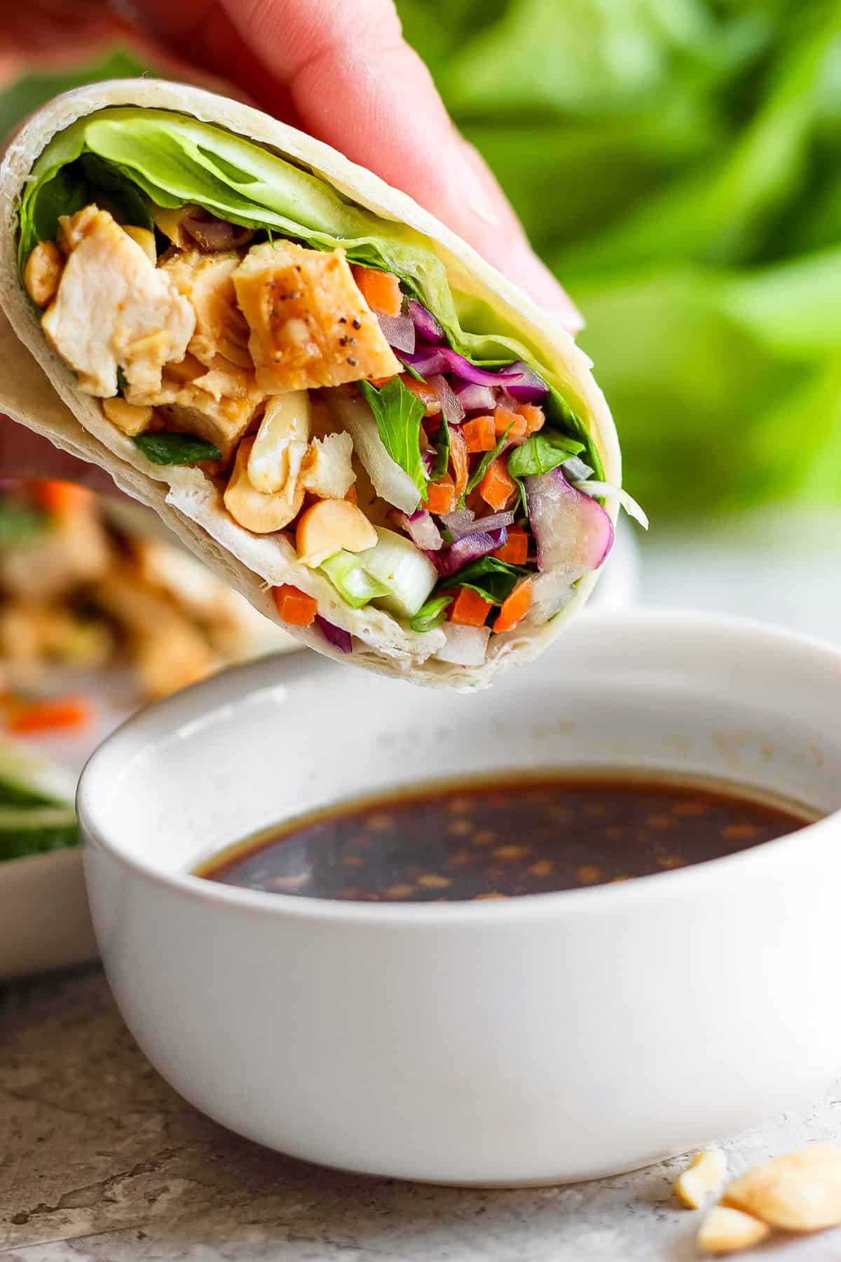 A hand holding a fresh spring roll filled with chicken, vegetables, and cashews, next to a bowl of dipping sauce.