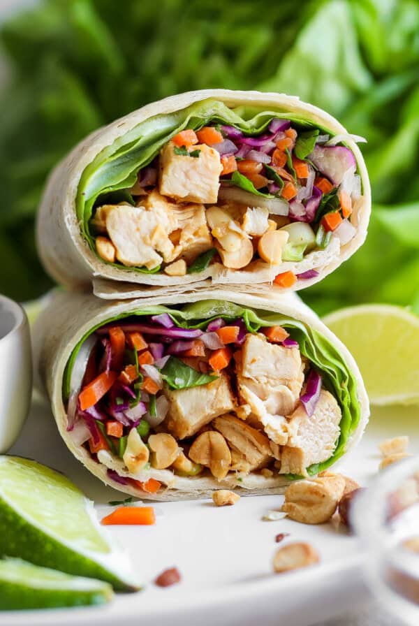 A fresh chicken wrap sliced in half, filled with mixed vegetables, lettuce, and peanuts, displayed with lime wedges and a salad in the background.