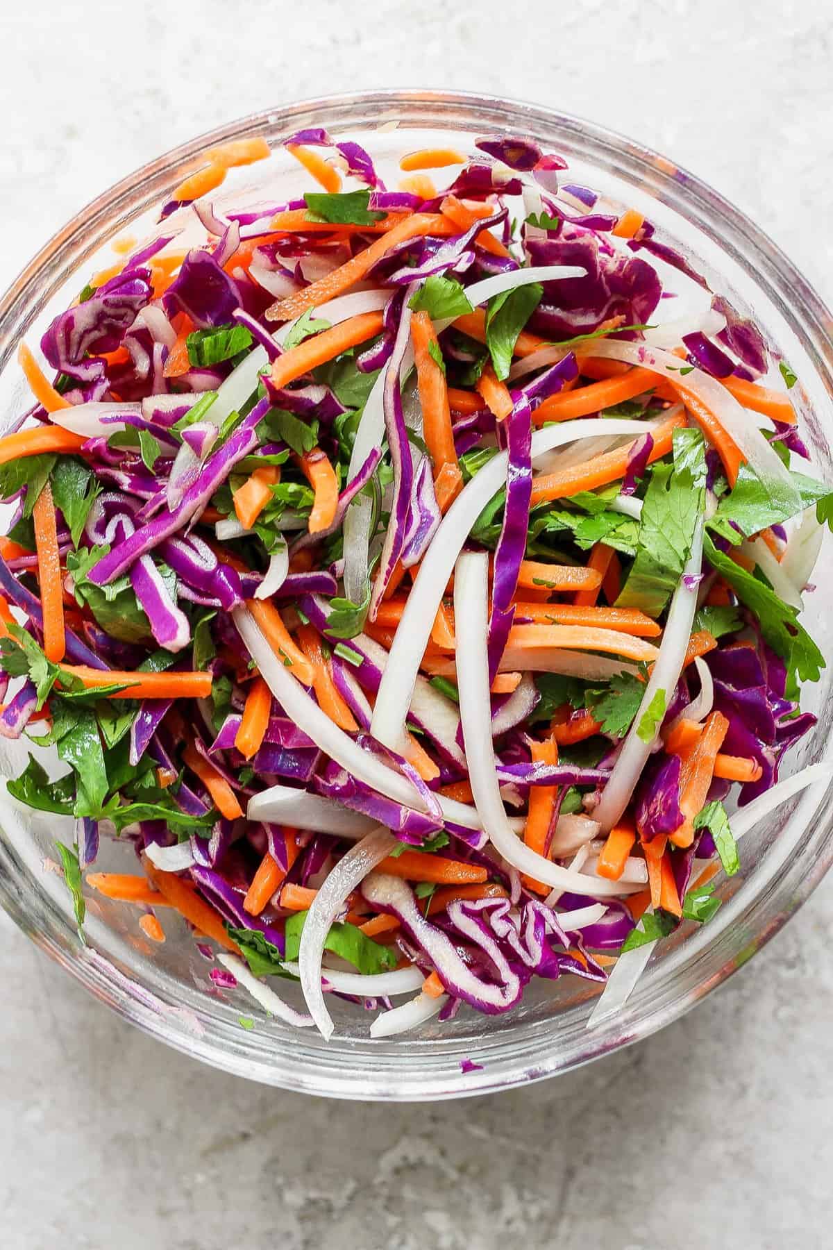 A colorful salad with red cabbage, carrots, onions, and parsley in a clear bowl on a white background.
