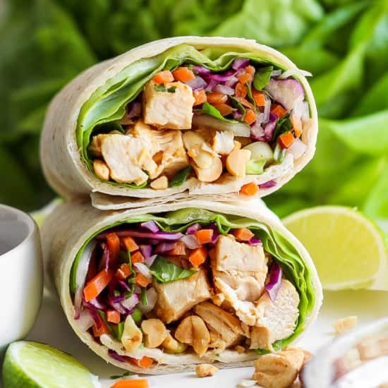 A close-up of a chicken wrap sliced in half, filled with diced chicken, purple cabbage, carrots, lettuce, and peanuts, displayed against a fresh, green backdrop.