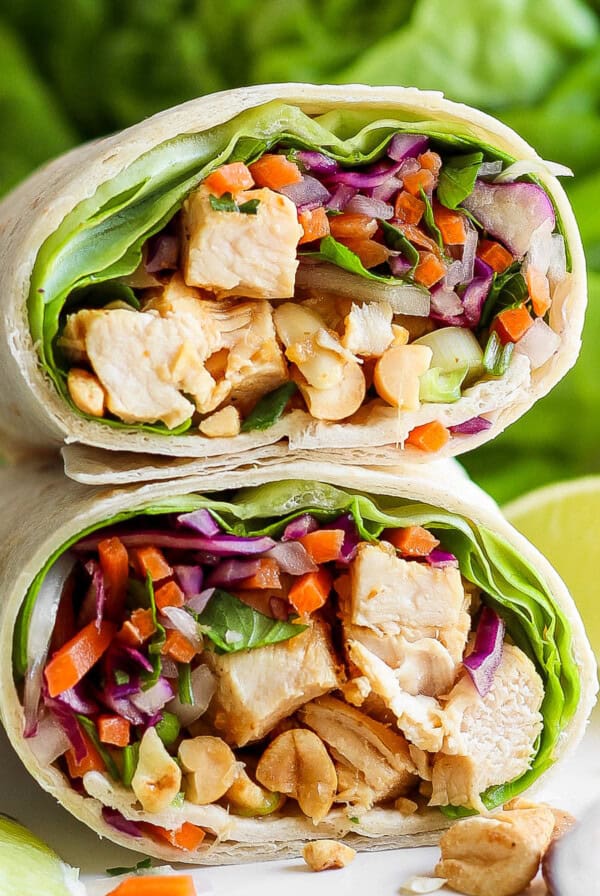 A close-up of a chicken wrap sliced in half, filled with diced chicken, purple cabbage, carrots, lettuce, and peanuts, displayed against a fresh, green backdrop.