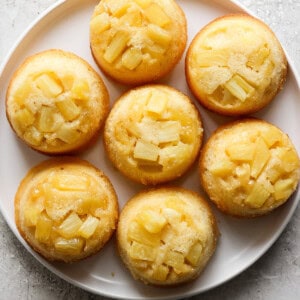 Plate of pineapple upside-down mini cakes topped with pineapple chunks on a light textured surface.