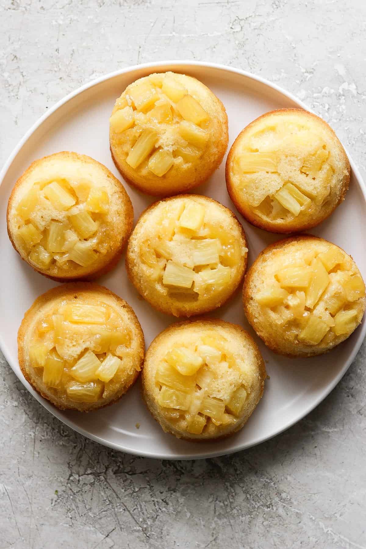 Plate of pineapple upside-down mini cakes topped with pineapple chunks on a light textured surface.