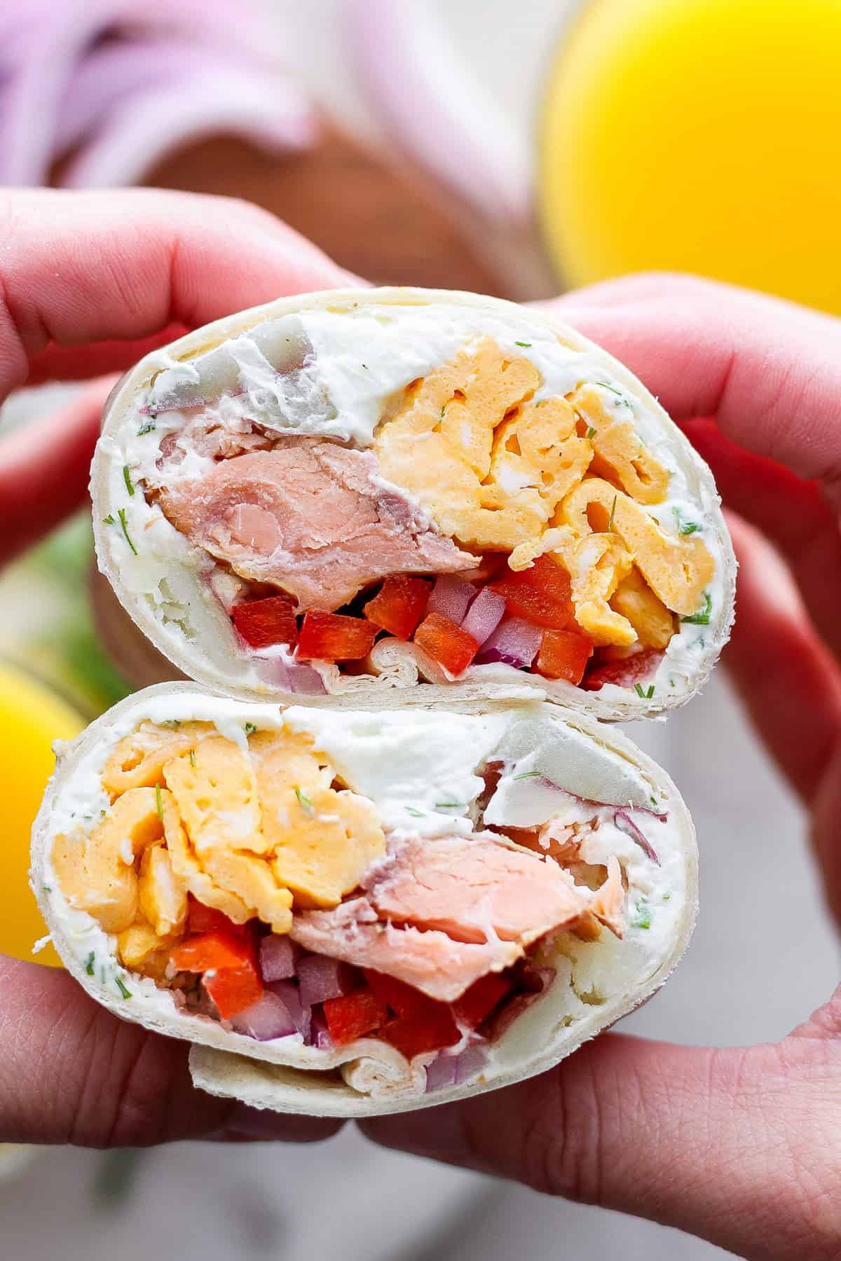 Close-up of a hand holding a halved wrap filled with salmon, cream cheese, diced vegetables, and scrambled eggs.