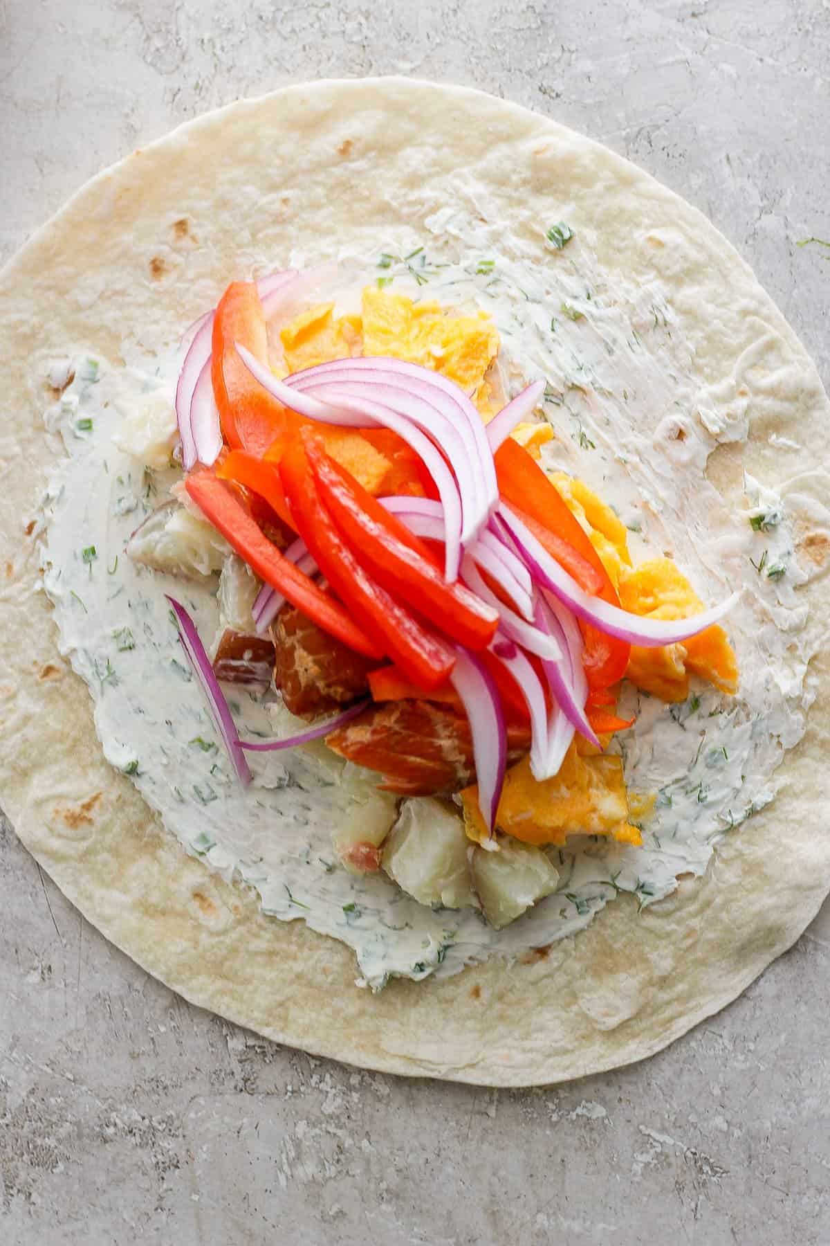A flour tortilla spread with herb cream cheese topped with slices of red bell pepper, red onion, and c،ks of chicken and orange.