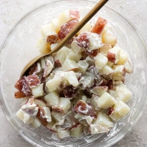 A bowl of potato salad with diced potatoes, creamy dressing, and bits of red onion, served with a wooden spoon on a light stone surface.