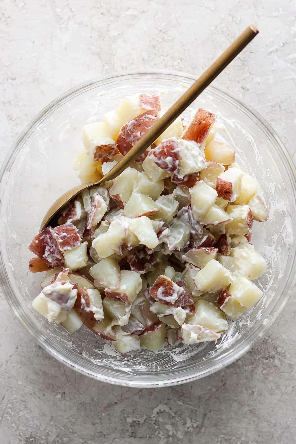 A bowl of potato salad with diced potatoes, creamy dressing, and bits of red onion, served with a wooden spoon on a light stone surface.
