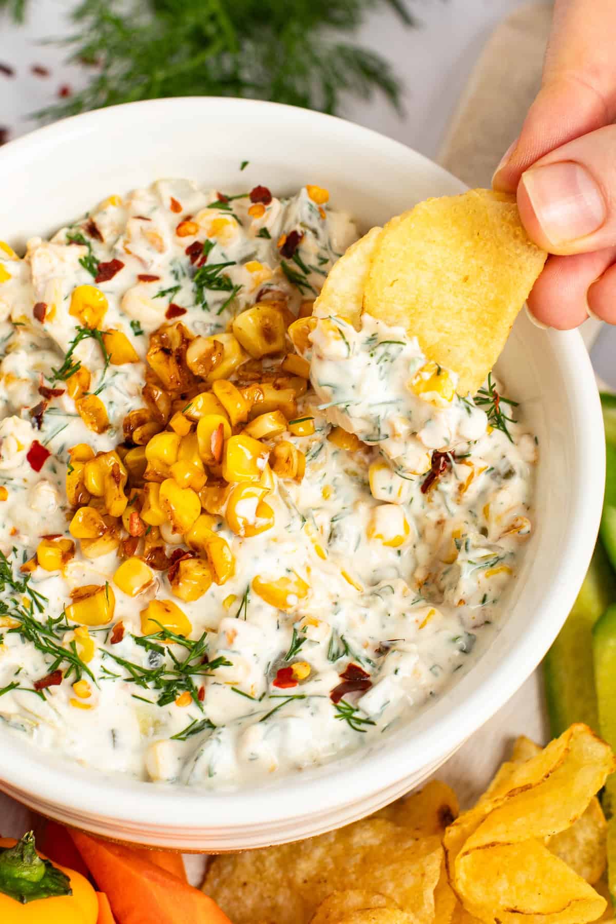 A hand dips a chip into a bowl of creamy dip topped with corn kernels and herbs, surrounded by chips and vegetable slices.