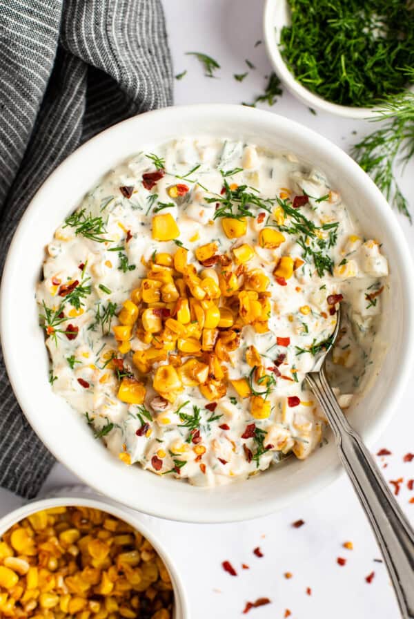 A bowl of creamy corn salad garnished with dill, red pepper flakes, and roasted corn kernels, with a spoon resting in it. Surrounding the bowl are a striped cloth and small bowls of dill and roasted corn.