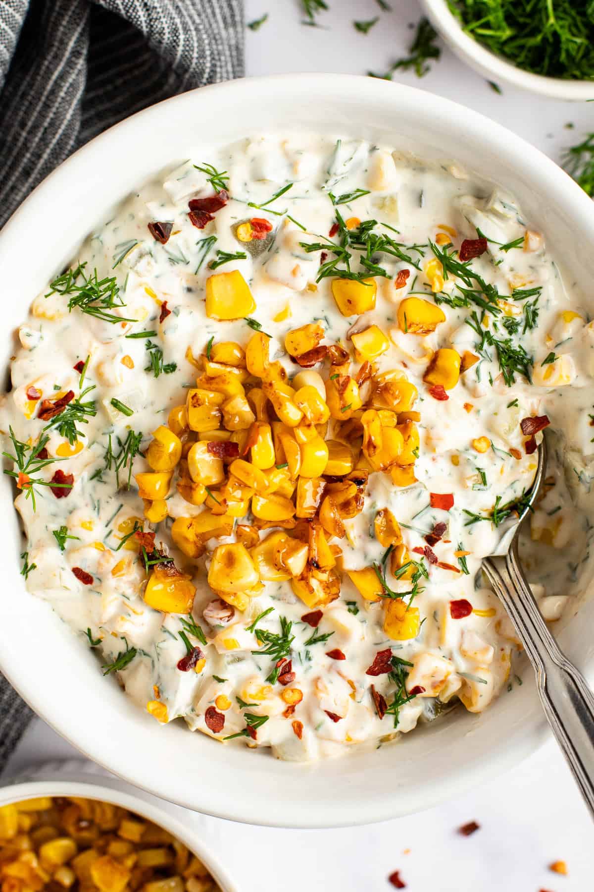 A bowl of creamy corn dip garnished with grilled corn, fresh herbs, and red pepper flakes, with a spoon resting inside the bowl.