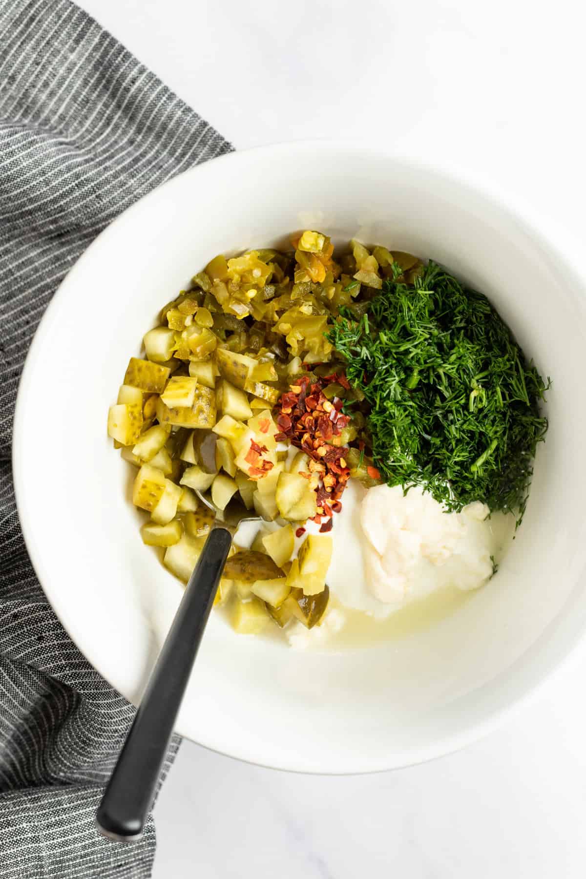 A white bowl holds chopped pickles, fresh herbs, crushed red pepper, and a dollop of creamy white sauce, with a black spoon resting inside. A gray-striped cloth is partially visible on the side.