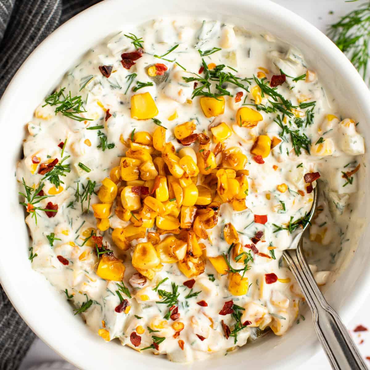 A bowl of creamy dip topped with roasted corn, chopped herbs, and red pepper flakes, with a spoon on the side.