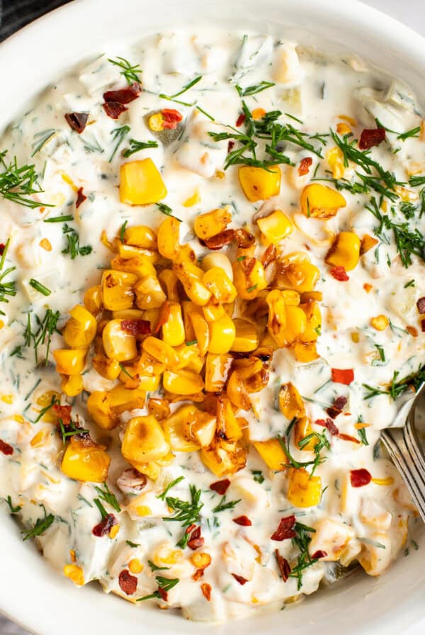 A bowl of creamy dip topped with roasted corn, chopped herbs, and red pepper flakes, with a spoon on the side.