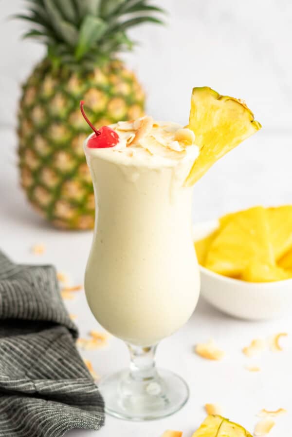 A tall glass of creamy Pina Colada garnished with a pineapple slice, cherry, and shredded coconut, with a whole pineapple and bowl of pineapple chunks in the background.