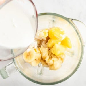 A measuring cup pours milk into a blender containing sliced bananas and pineapple chunks.