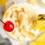 Top view of a creamy tropical cocktail garnished with toasted coconut flakes, a maraschino cherry, and a pineapple slice.