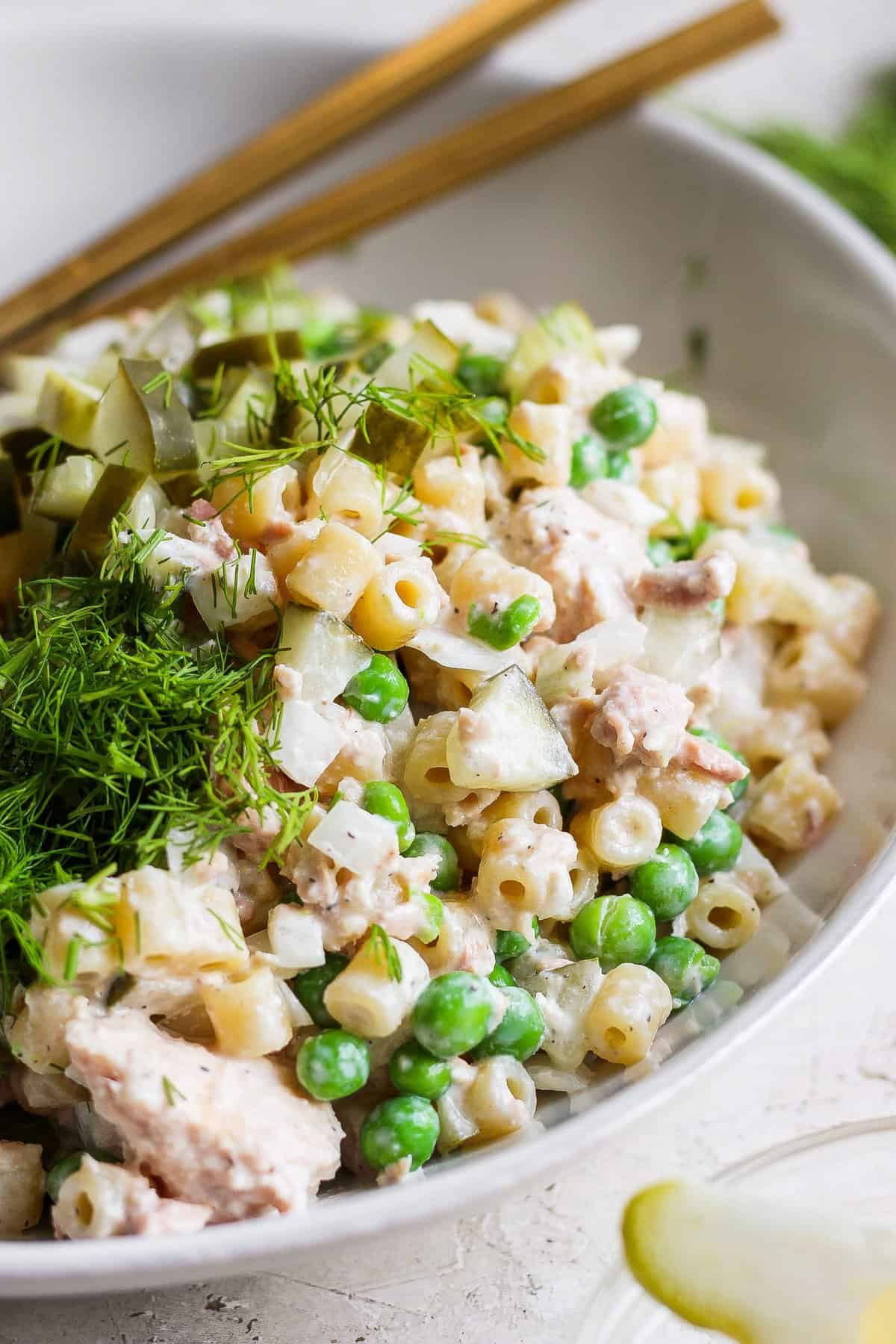 A bowl of tuna pasta salad with peas and dill, garnished with fresh herbs and served with c،psticks.