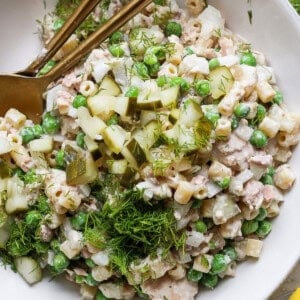 A bowl of tuna salad with peas, chopped cucumbers, and dill, served with a pair of gold chopsticks on a white plate.