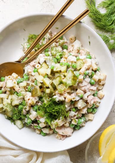 A bowl of tuna salad with peas, chopped cucumber, and dill, served with a pair of chopsticks and fresh lemon slices on a neutral-toned background.