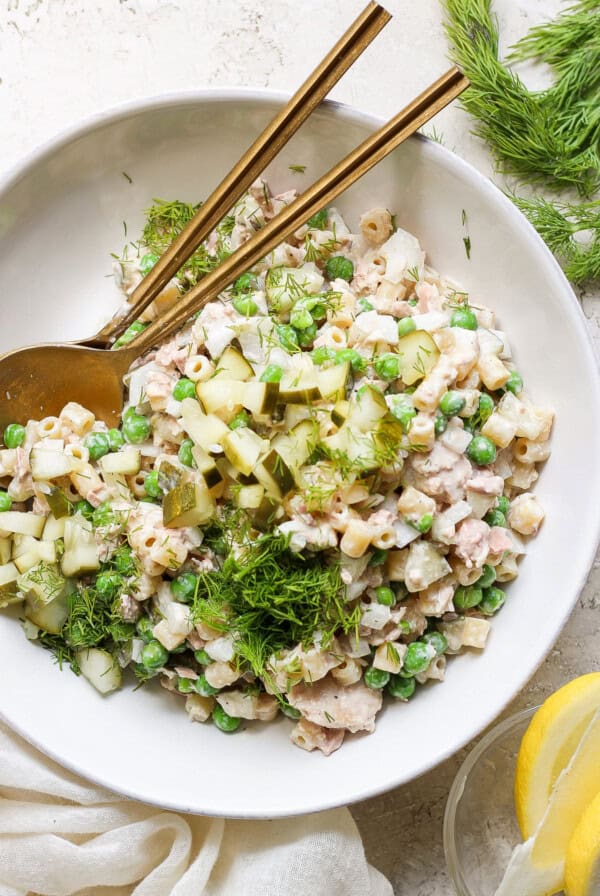 A bowl of tuna salad with peas, chopped cucumber, and dill, served with a pair of chopsticks and fresh lemon slices on a neutral-toned background.