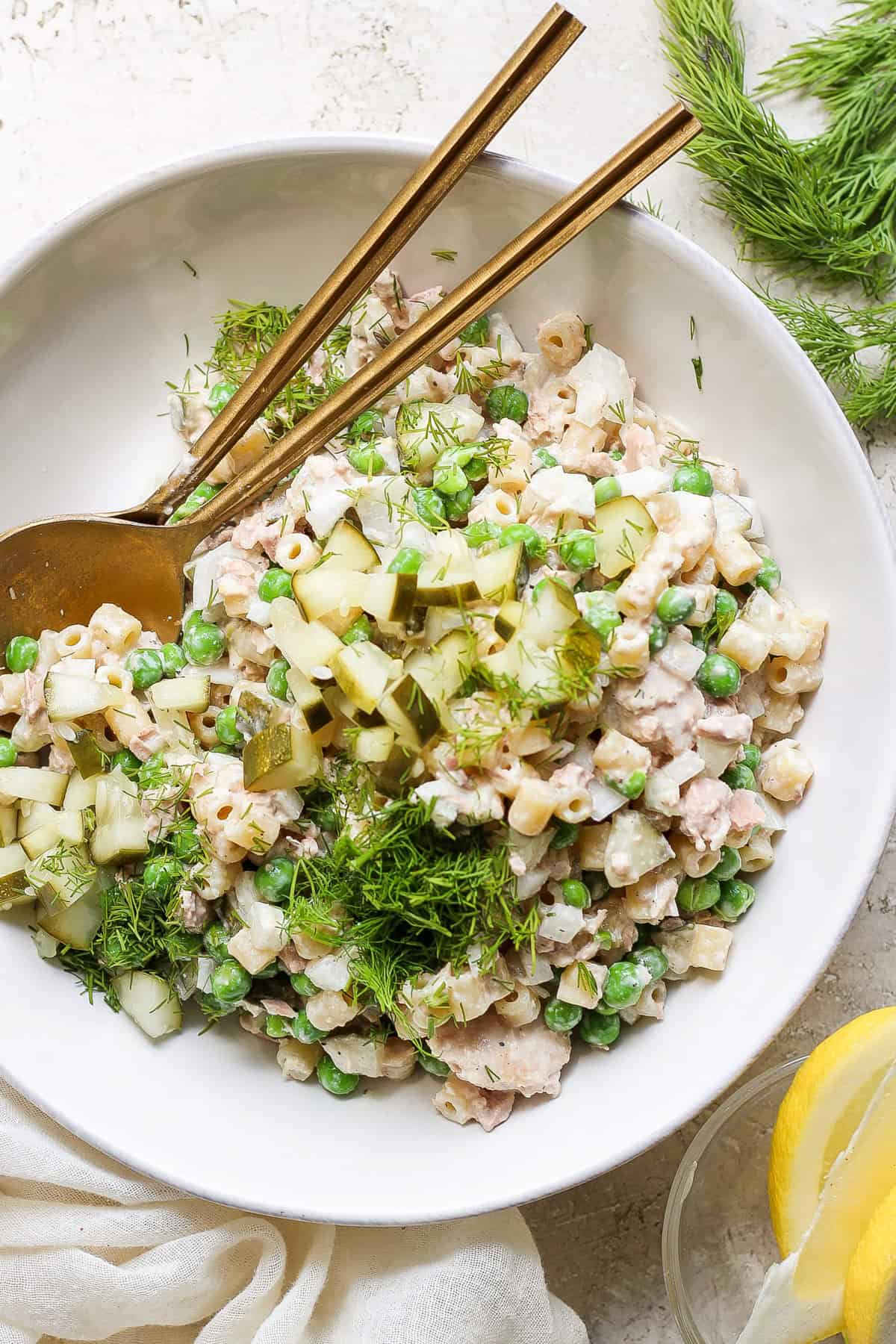 A bowl of tuna salad with peas, c،pped cu،ber, and dill, served with a pair of c،psticks and fresh lemon slices on a neutral-toned background.