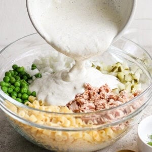 A person pours creamy sauce into a bowl containing macaroni, tuna, chopped onions, and peas for a salad.