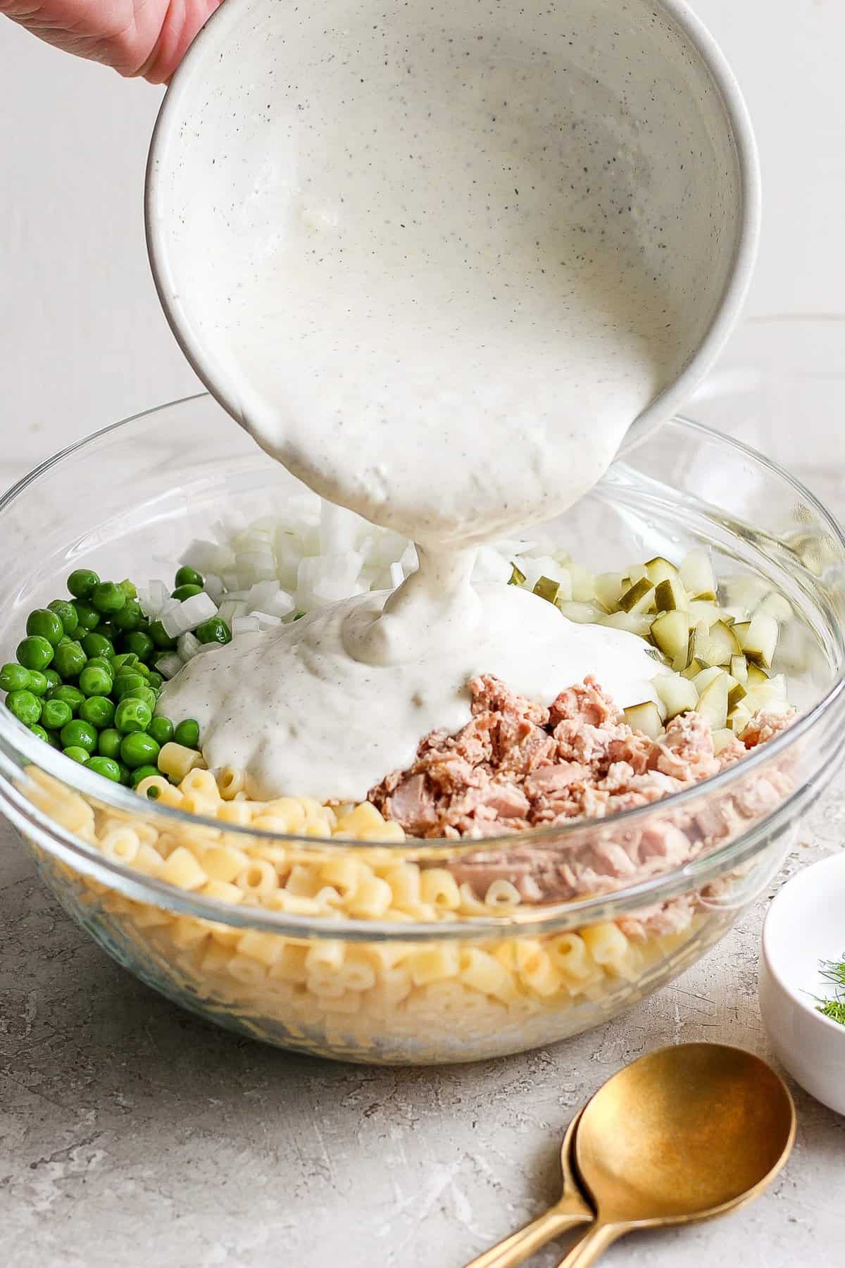 A person pours creamy sauce into a bowl containing macaroni, tuna, chopped onions, and peas for a salad.