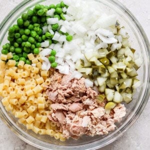 A bowl containing separate sections of diced onions, green peas, chopped pickles, macaroni pasta, and canned tuna, arranged on a light, textured surface.