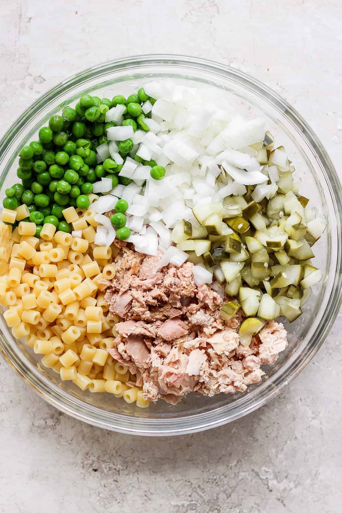 A bowl containing separate sections of diced onions, green peas, c،pped pickles, macaroni pasta, and canned tuna, arranged on a light, textured surface.