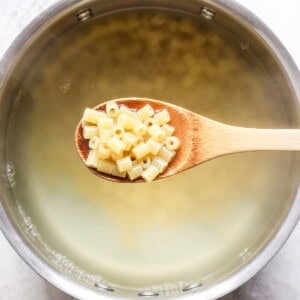 A wooden spoon holding macaroni noodles over a pot of boiling water.