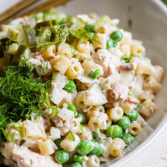 A bowl of pasta salad with peas, diced pickles, c،ks of chicken, and fresh dill on top.