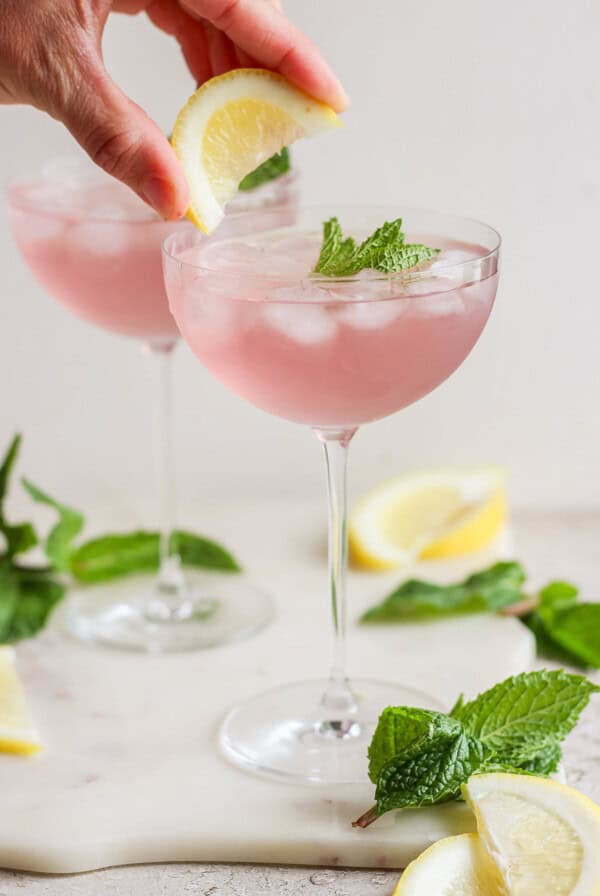 A person's hand placing a lemon wedge on the rim of a glass filled with pink lemonade garnished with mint leaves, with another similar glass in the background.