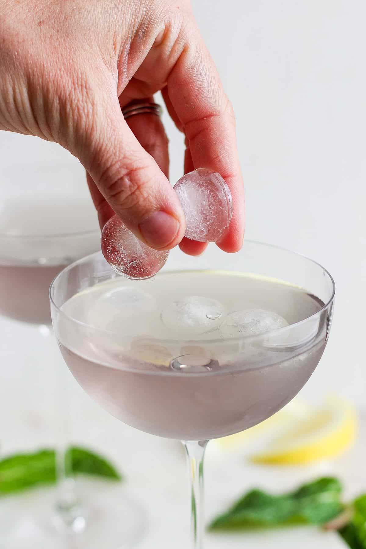 A hand placing ice cubes into a cocktail glass filled with a clear drink, with more ice-filled glasses and lemon slices blurred in the background.