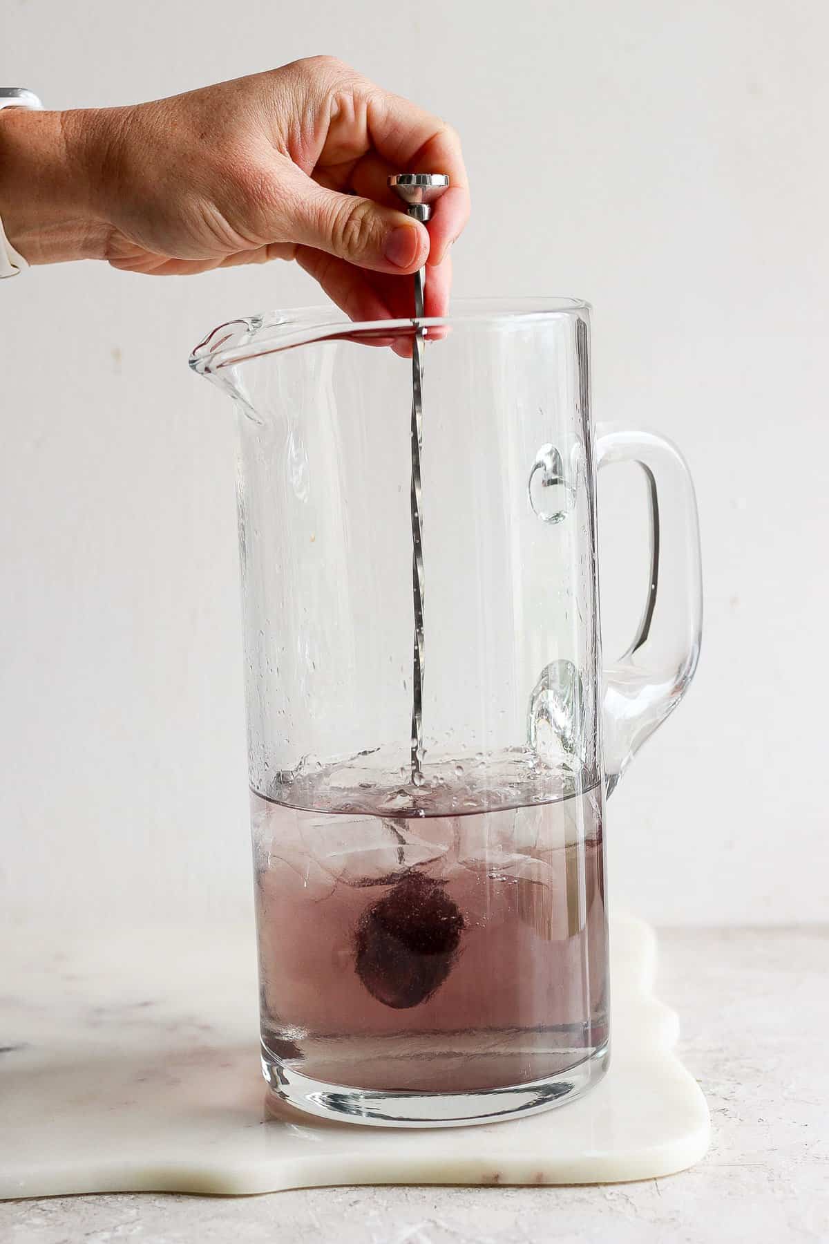 A hand uses a metal spoon to stir a dissolving purple effervescent tablet in a clear glass pitcher filled with water.