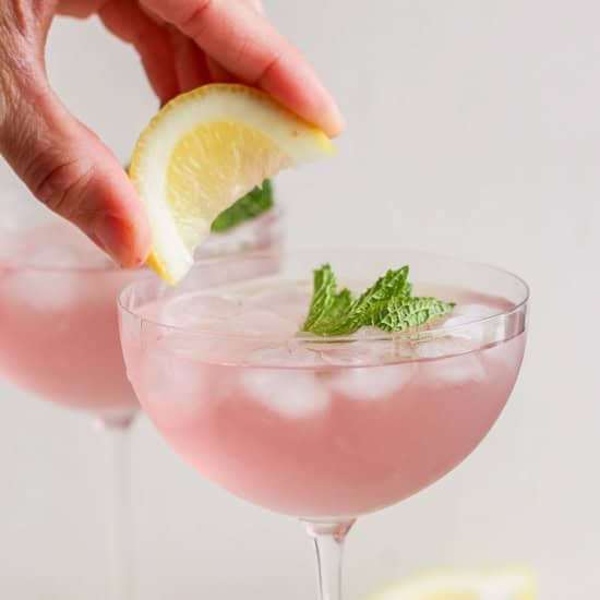 A hand placing a lemon slice on the rim of a pink cocktail garnished with mint in a stemmed glass.
