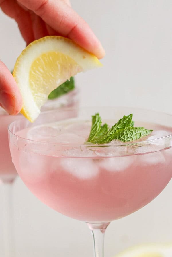 A hand placing a lemon slice on the rim of a pink cocktail garnished with mint in a stemmed glass.
