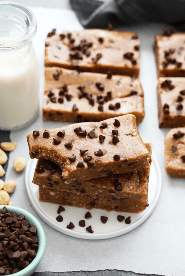 A stack of chocolate chip cookie dough protein bars, reminiscent of homemade larabars, on a white plate surrounded by additional bars, a glass of milk, chocolate chips, and cashews.