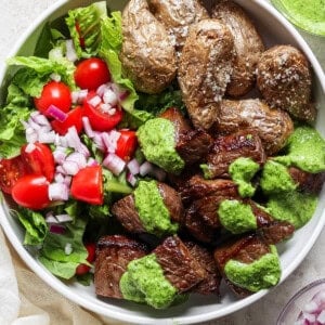 A bowl of grilled steak chunks with green sauce, roasted potatoes, and a side salad of lettuce, tomatoes, and chopped red onions.