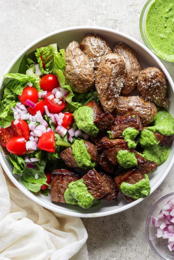 A bowl of grilled steak chunks with green sauce, roasted potatoes, and a side salad of lettuce, tomatoes, and chopped red onions.